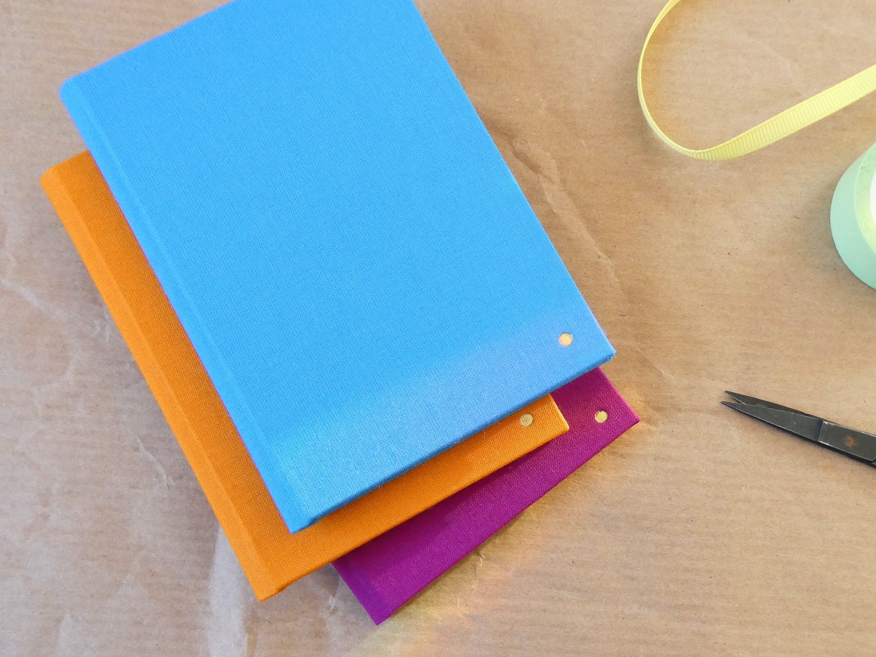 Three colourful notebooks stacked on a brown paper surface with scissors and a roll of tape.
