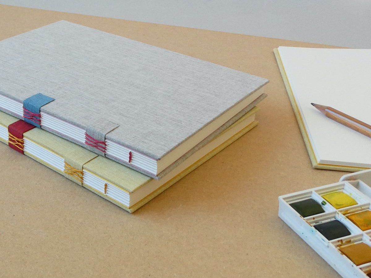 Stack of notebooks with colourful binding options on a beige surface, with a watercolor set in the foreground.