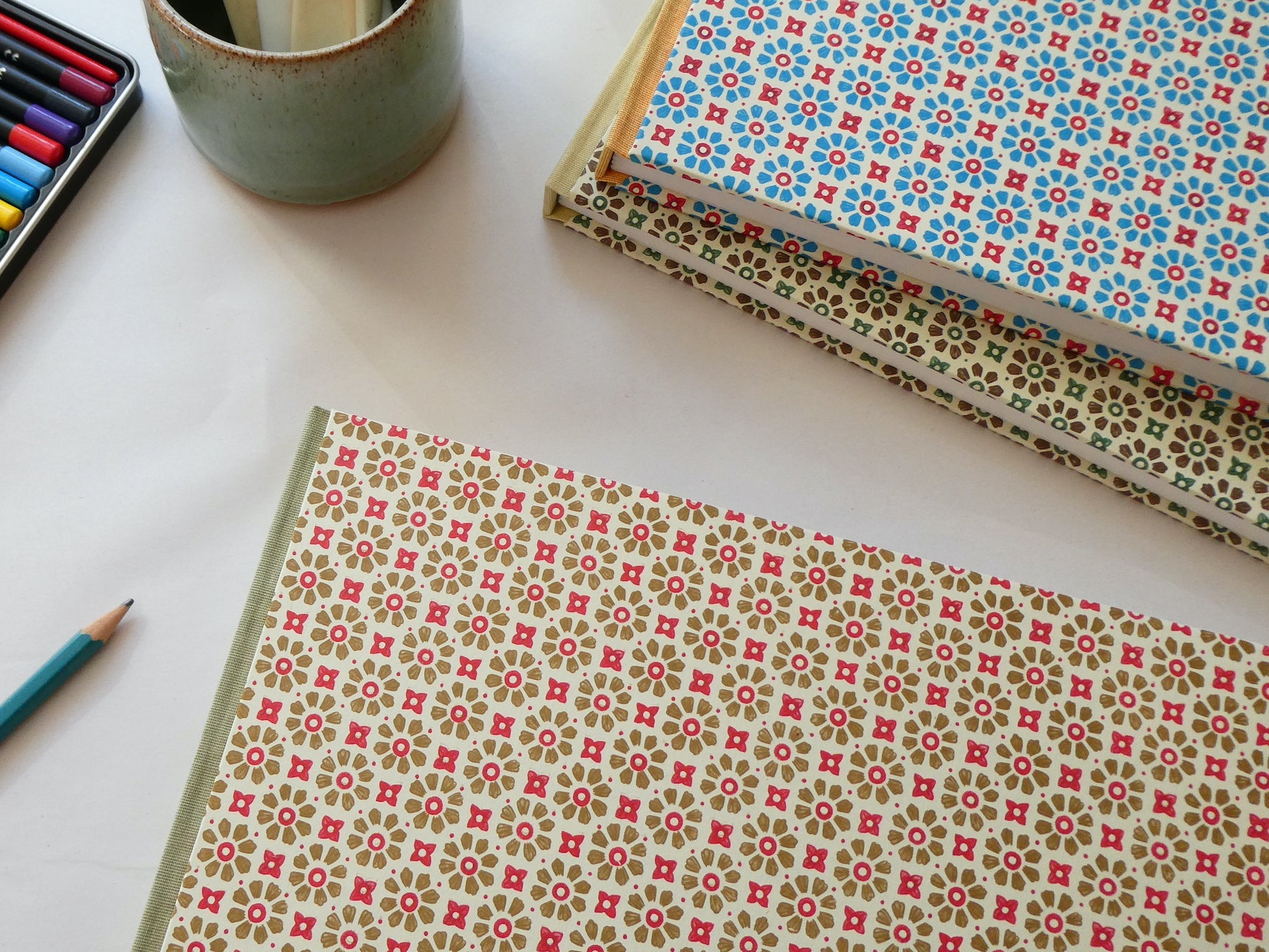 Three hand bound watercolour sketchbooks with Italian decorative paper covers on a white surface with mug and pencils