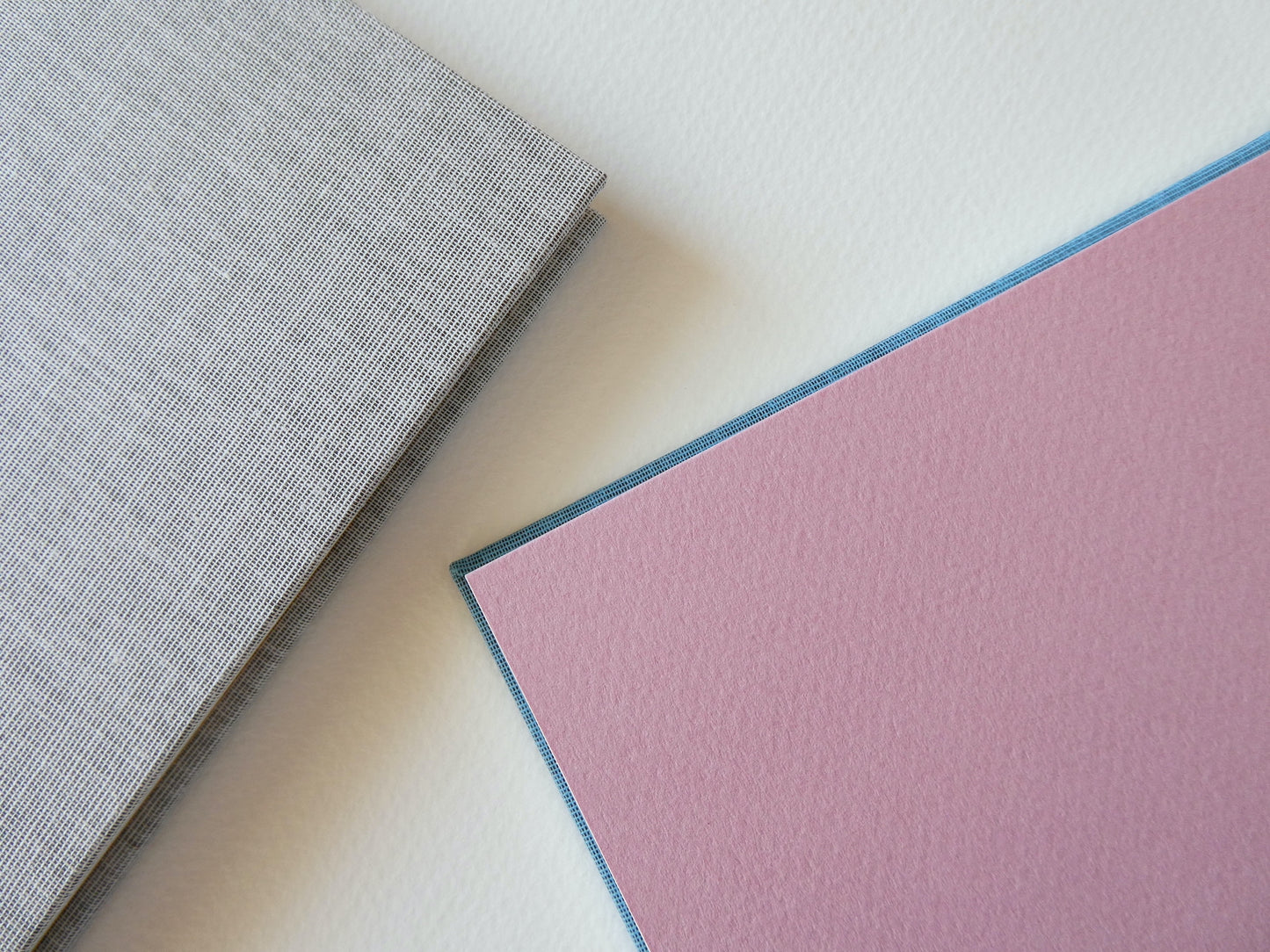 Close up of two hand bound sketchbooks, showing pink endpapers and grey bookcloth, on a light surface.
