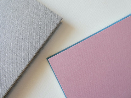 Close up of two hand bound sketchbooks, showing pink endpapers and grey bookcloth, on a light surface.